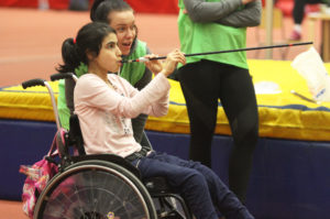 Girl in a wheelchair plays with a blowpipe, assisted by an adult wearing a green bib